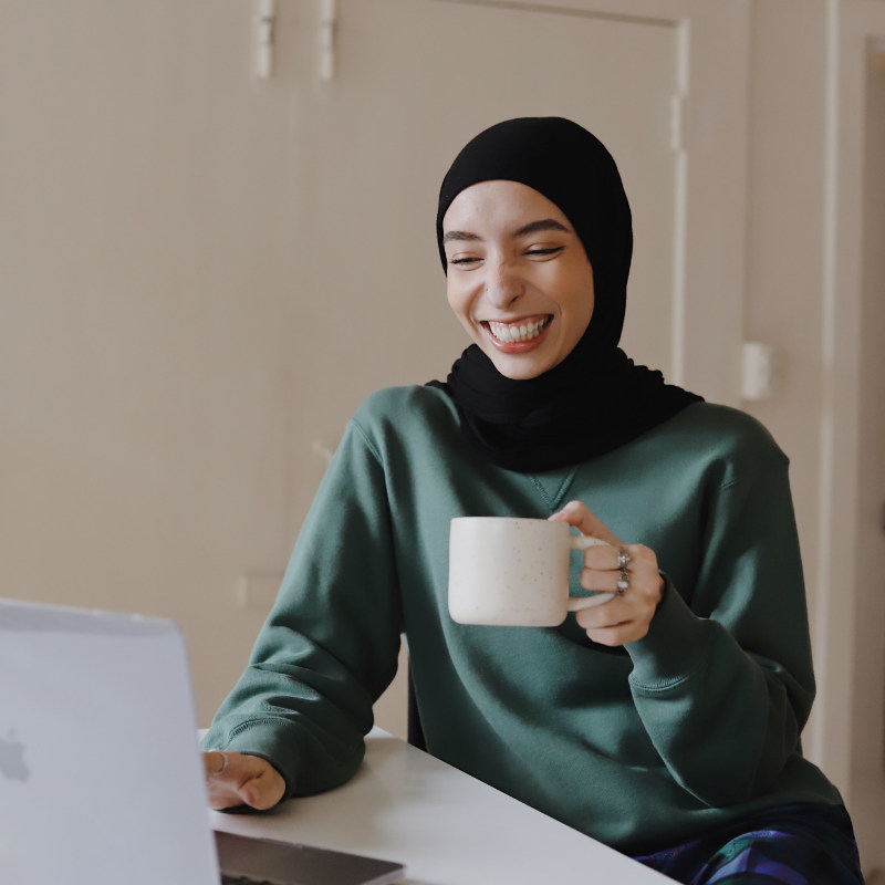 Image of a woman smiling while sitting at a desk, holding a white cup and looking at a portable computer. The woman is wearing a green sweater and a black hijab. She is in a room with white walls and doors.